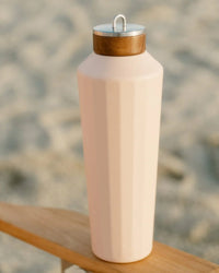 The 25oz Hemingway Flask by Business & Pleasure Co in Dusty Pink, featuring a wooden-look cap and vacuum insulation to keep drinks fresh for hours, rests on a wooden surface with blurred sand in the background.