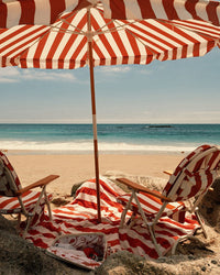 Two Crew Le Sirenuse Stripe beach chairs and a matching Amalfi Umbrella by Business & Pleasure Co face the ocean on sandy shores, with a red-and-white towel and beach bag beneath, set against blue skies and rolling waves.