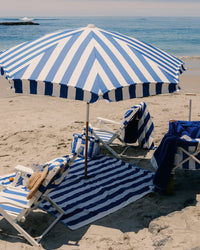 The Amalfi Umbrella - Crew Mediterranean Blue Stripe by Business & Pleasure Co shades two matching lounge chairs and a towel on the sand, with beach accessories, including a hat, arranged beneath this stylish sun shelter on the shore.