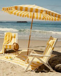 Two yellow and white striped beach chairs and a Business & Pleasure Co Amalfi Umbrella - Crew Mimosa Yellow Stripe sit by the ocean. A towel, sunhat, and sunglasses rest on the chairs—enjoy perfect shade under this UV-resistant umbrella.
