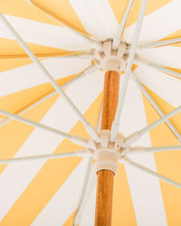 Close-up of the Amalfi Umbrella - Crew Mimosa Yellow Stripe by Business & Pleasure Co, showing its yellow and white UV-resistant canopy, wooden pole, and white spokes radiating outward.