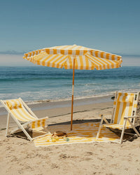 Two yellow-and-white beach chairs and a Business & Pleasure Co Amalfi Umbrella - Crew Mimosa Yellow Stripe are set up on sand facing the ocean, with a matching towel, sandals, and a drink below a clear blue sky.