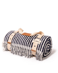 The Beach Blanket - Laurens Navy Stripe by Business & Pleasure Co is a rolled-up, 100% cotton striped blanket with fringe edges, secured by beige straps with leather accents, shown on a white background.