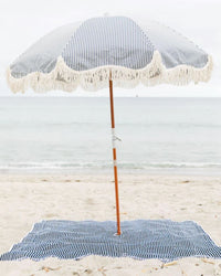 A blue-and-white striped beach umbrella with fringe shades the Business & Pleasure Co Beach Blanket - Laurens Navy Stripe on sand, set against calm ocean water and a cloudy sky.