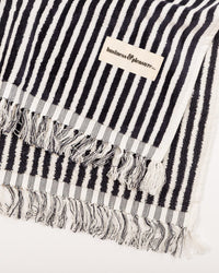 The Beach Blanket - Laurens Navy Stripe by Business & Pleasure Co is a family-friendly, black and white striped, 100% cotton blanket with fringed edges and a sewn-on brand label.