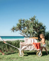 Two children push a Business & Pleasure Co Beach Cart - Laurens Pink Stripe, loaded with orange towels, on grass near the beach, with trees, ocean, and a clear blue sky in the background.