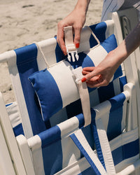 Close-up of hands attaching a beige strap to the Tommy Chair - Acacia White - Crew Mediterranean Blue Stripe by Business & Pleasure Co, featuring a matching padded headrest and set on sandy ground.