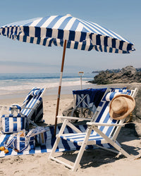 A beach scene with a Business & Pleasure Co Tommy Chair in Acacia White and Crew Mediterranean Blue Stripe, plus an umbrella, towel, bags, and a straw hat on the sand. The ocean, sky, and shoreline rocks are in the background.