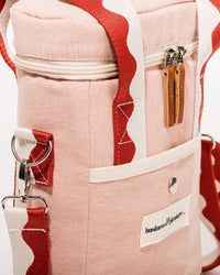 A close-up of the Business & Pleasure Co Wine Cooler Tote Bag - Rivie Pink, featuring vegan leather, a red and white strap, front snap pocket, two zipper pulls, and a metal ring attachment.