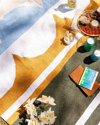 A vibrant picnic unfolds on the Luna Lago - Blanket Roll by caminito, featuring drinks, snacks, a book, notebook, fresh flowers in a jar, and soda cans—sunlit and secured with a handmade leather strap around the lightweight blanket.