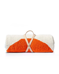 The caminito Luna Paseo - Blanket Roll is a lightweight, textured outdoor blanket in orange and white, rolled up and secured with a handmade leather strap and handle, shown against a plain white background.