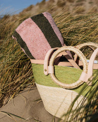 A woven straw bag with circular handles rests on the sand in beach grass, carrying the caminito Marea Fresca - Blanket Roll in pink and dark green stripes for a relaxed, outdoor beach vibe.