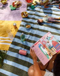 A person sits on the caminito Marea Lago - Blanket Roll, handwoven in Mexico, reading a colorful magazine. Drinks, snacks, and a doll are nearby, with another blanket holding more food and a childs legs seen in the background.