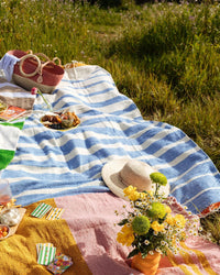 A summer picnic scene with the caminito Marea Paseo - Blanket Roll spread on the grass, a straw hat, yellow flowers in a vase, a basket, food, drinks, and playing cards scattered in the sunlight.