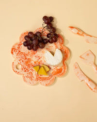 A Casa Amarosa 9in Scallop Resin Cheese/Charcuterie Board in Ivory Pink displays red grapes, a wheel of white cheese, and two yellow cheese slices. Matching marbled cheese utensils are arranged on the right.