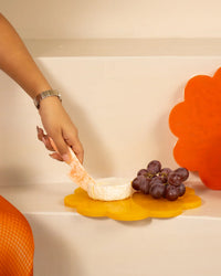 A hand with a cheese knife hovers over cheese and grapes on the Casa Amarosa 9in Scallop Resin Cheese/Charcuterie Board in Mustard, shaped like a yellow flower.