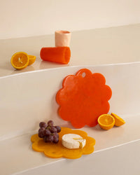 A Casa Amarosa 9in Scallop Resin Cheese/Charcuterie Board in Orange, an orange cup, and half an orange sit above a yellow handmade cheese board with grapes, cheese, and orange halves.