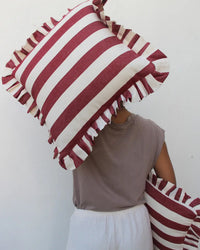 A person in a taupe shirt and white pants holds two Casa Amarosa Cherry Ruffle 16x16in pillows—one covering their face, the other by their side—against a plain white background.