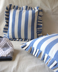 Two Casa Amarosa Denim Ruffle 16x16in pillows with blue and white stripes and ruffled edges rest on a light fabric surface, accompanied by a textured, folded blue-and-white patterned cloth nearby.