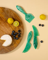 Round wooden charcuterie board with brie, blueberries, a lemon, a halved lime, and three Casa Amarosa Resin Cheese Knives in Green displayed on a beige surface.