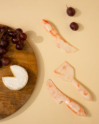 The Casa Amarosa Resin Cheese Knife Set of 3 in Ivory Pink is displayed on a beige surface beside red grapes and a round of brie on a wooden board.