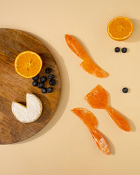 A wooden board with cheese, blueberries, and a halved orange sits next to the Casa Amarosa Resin Cheese Knife Set of 3 in Orange and more blueberries on a beige surface.