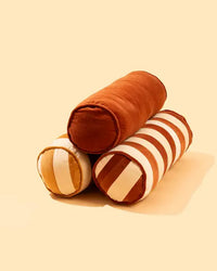 Three Casa Amarosa Velvet Stripe Bolster Pillows are stacked on a plain beige background: one in solid rust, one in solid beige, and one with clay and off-white stripes—ideal for enhancing your home décor.