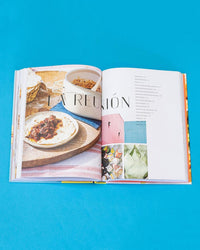 An open copy of Ama by Chronicle Books shows a tortilla dish photo on the left and a table of contents with images on the right, set against a blue background—perfect for discovering Tex-Mex recipes and regional Mexican cuisine.
