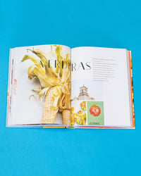 An open book, Ama by Chronicle Books, rests on a blue surface showing images of corn, a building, and a tomato with VERDURAS in Spanish—hinting at Tex-Mex recipes inspired by regional Mexican cuisine.