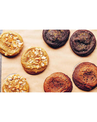 Six round cookies—three light brown, three chocolate-colored, some topped with almonds—are arranged on parchment paper, offering tasty inspiration for vegan or gluten-free treats from Cookies & Crumbs by Chronicle Books.