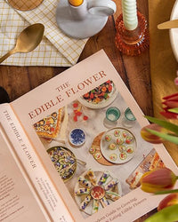 An open book titled Edible Flower by Chronicle Books shows dishes with edible flowers on the cover, placed among tableware, a candle, and fresh flowers on a wooden table.