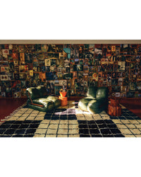 Two green lounge chairs and small wooden tables rest on a checkered rug beside Flamingo Estate: The Guide to Becoming Alive by Chronicle Books, evoking nature-inspired wellness, with photos and art displayed on the wall behind.