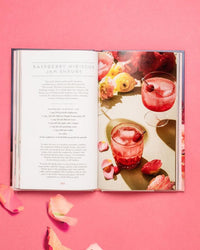 An open copy of Floral Libations by Chronicle Books rests on a pink surface, displaying a Raspberry Hibiscus Jam Shrubs recipe with two pink drinks and flowers—perfect inspiration for floral cocktails and edible flower drinks.