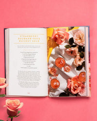 The open Floral Libations cocktail book by Chronicle Books rests on a pink background, showing a strawberry rhubarb-rose whiskey sour recipe and a photo of floral drinks surrounded by roses.