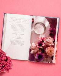 The open book Floral Libations by Chronicle Books rests on a pink surface, displaying the “Dandelion Honey Bowl of Soul” recipe and an elegant floral cocktail photo surrounded by pink blossoms—ideal for edible flower cocktail fans.