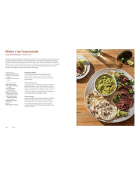 A plate with grilled steak slices, guacamole, lime wedges, and warm tortillas sits beside salsa on a wooden table, inspired by family recipes from Mamacita by Chronicle Books.