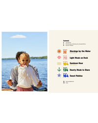 A toddler in a white sweater stands by an adult at a lake; next to them is the Sea Lovers Cookbook by Chronicle Books, open to a colorful contents page that features sections on sustainable cooking and pescatarian recipes.