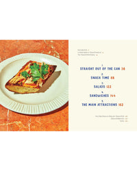 A slice of toast with tinned fish and herbs rests on a plate beside the Tin to Table cookbook from Chronicle Books, open to a bold, colorful contents page highlighting its tinned seafood recipe sections.