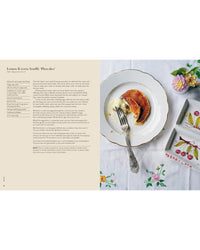 A plate with a partially eaten lemon ricotta soufflé pancake, a fork, and a dish labeled MARMELLATA sits on an embroidered tablecloth next to Wild Figs and Fennel by Chronicle Books.