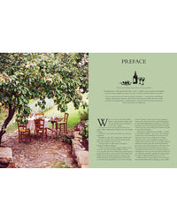 A rustic outdoor dining set sits on a stone patio beneath a leafy tree, beside Chronicle Books’ “Wild Figs and Fennel,” Letitia Clark’s Mediterranean cookbook with Italian recipes illustrated with wine, cheese, and grapes.