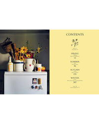 A white fridge with magnets stands in a kitchen beside a counter with a vase of yellow flowers, ceramic jug, bottle, and fruit; on the right is a contents page from Chronicle Books’ Wild Figs and Fennel cookbook.