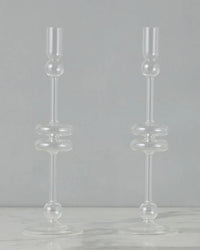The etúHOME Clear Glass Candlesticks Set of 2 - Large features modern, geometric round and cylindrical shapes, displayed on a light surface with a gray background.