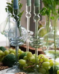 A close-up of etúHOMEs Clear Glass Candlesticks Set of 2 - Large on a table, surrounded by green grapes, limes, and fresh greenery.
