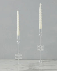 Two white twisted taper candles are displayed in the etúHOME Clear Glass Candlesticks Set of 2 - Large, handmade from recycled glass, on a white marble surface against a plain gray background.