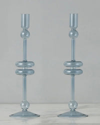 Two identical Denim Glass Candlesticks Set of 2 - Large by etúHOME, handmade from recycled glass with rounded, spherical accents, stand on a marble surface against a plain light background.