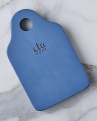 The etúHOME Dutch Small Cheese Board w/ Spreader in Denim Blue, featuring a handle and hole, sits on a white marble surface.