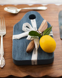The etúHOME Dutch Small Cheese Board w/ Spreader in Denim Blue is styled with a ribbon and lemon with leaves, displayed on a wooden table next to a fork, spoon, and knife.