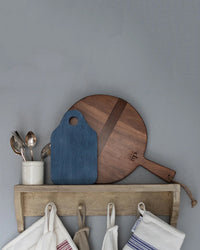 A wooden shelf displays the etúHOME Dutch Small Cheese Board w/ Spreader in Denim Blue, a cup with assorted spoons, and dish towels hanging from hooks underneath.
