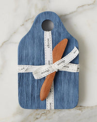 The etúHOME Dutch Small Cheese Board w/ Spreader in Denim Blue, tied with a white ribbon, sits atop a white marble surface.