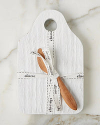 The etúHOME Dutch Small Cheese Board w/ Spreader in White features a textured board with a top hole, paired with a wooden spoon and spreader tied by a white ribbon, all displayed on marble.
